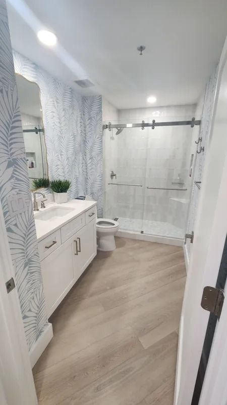 Bright modern bathroom with white vanity, toilet, and glass shower, light wood floor and patterned walls