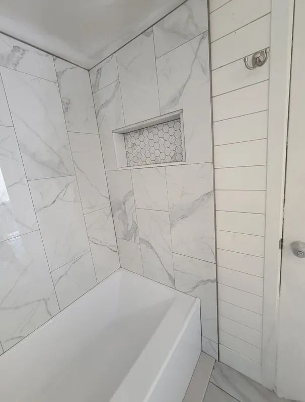 Small white tiled bathroom with a bathtub, recessed shelf, and showerhead.