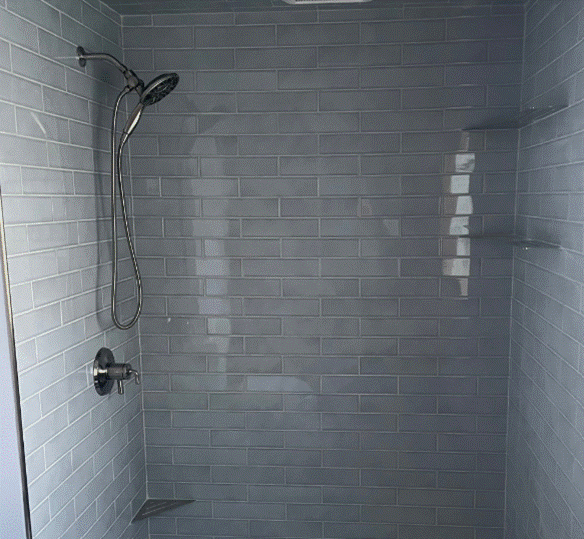 Gray tiled shower with a showerhead, shelf, and faucet.