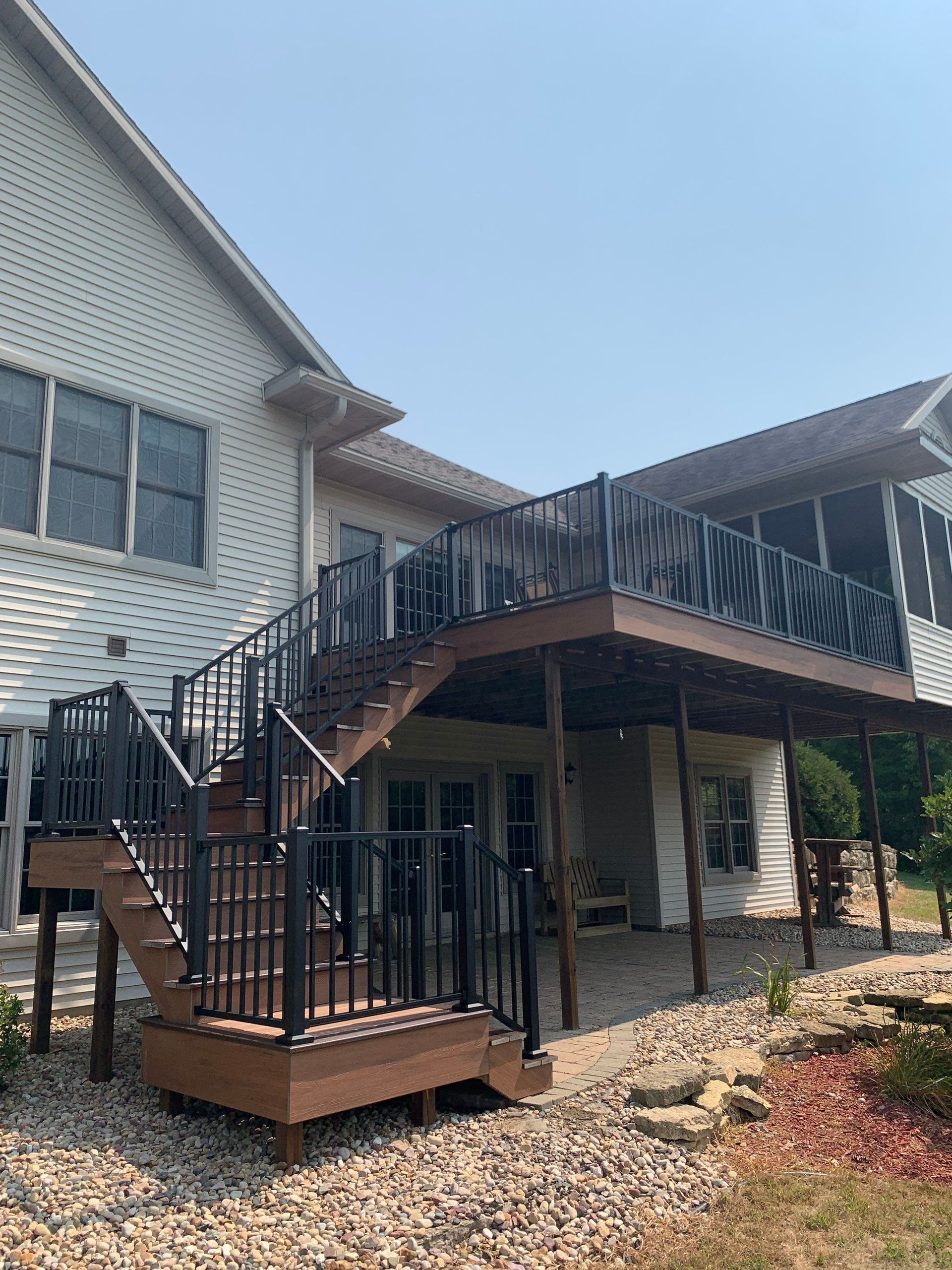 Brown deck with black railings attached to a two-story house, accessed by stairs.