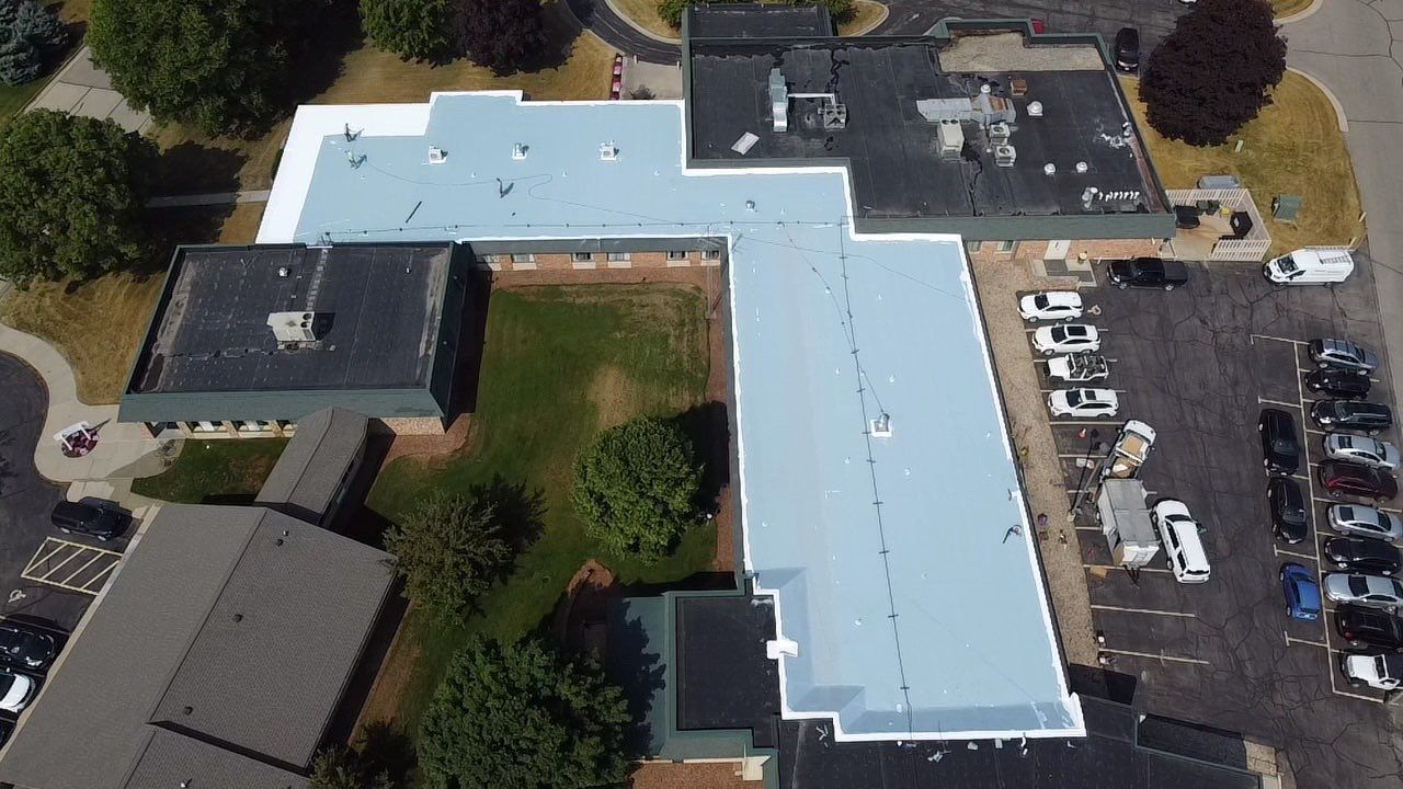 Aerial view of a building complex with multiple roofs, a parking lot with vehicles, and green space.