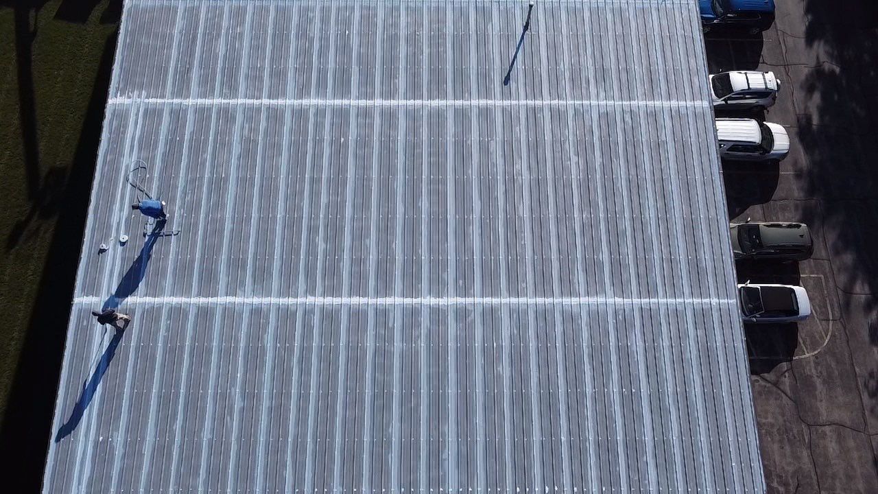Aerial view of a large, light-colored, corrugated roof with parked cars along the right side and figures on the left.