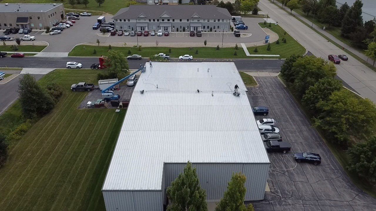 Aerial view: industrial building with metal roof and parked vehicles in a grassy lot, other buildings in the background.