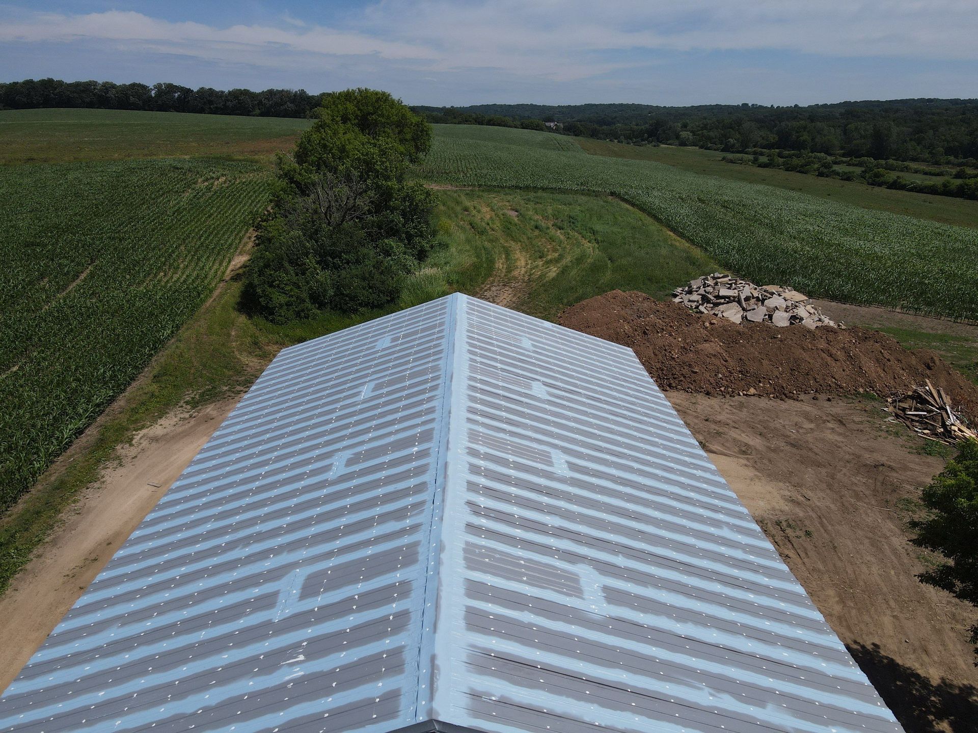 Aerial view of a metal roof on a barn in a field with green crops under a sunny sky.