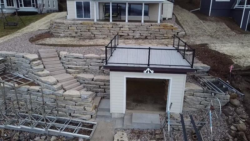 Stone retaining walls and stairs lead to a boathouse with a dock on a lake.