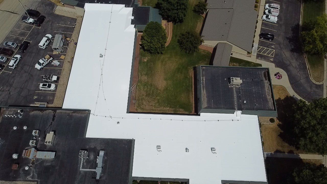 Aerial view of buildings with white and dark roofs, a parking lot with cars, and some trees.