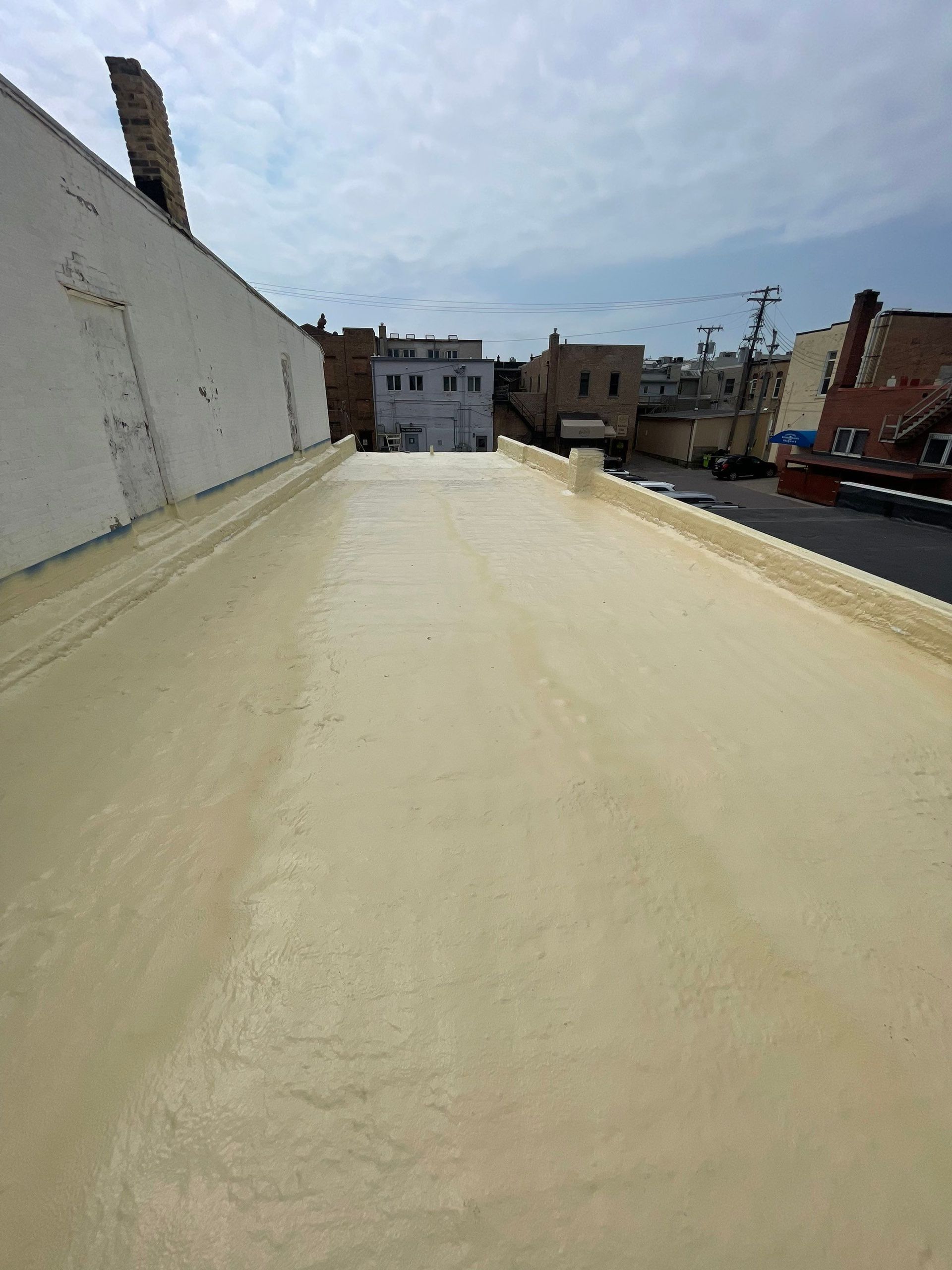 Flat roof covered in light tan foam with surrounding buildings and sky in background.