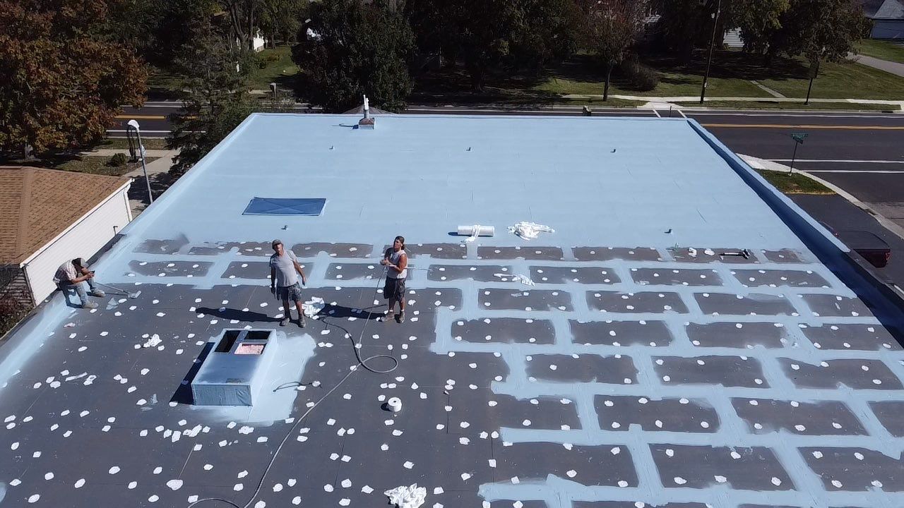 Construction workers on a flat, light blue roof; applying dark rectangles; sunny day.