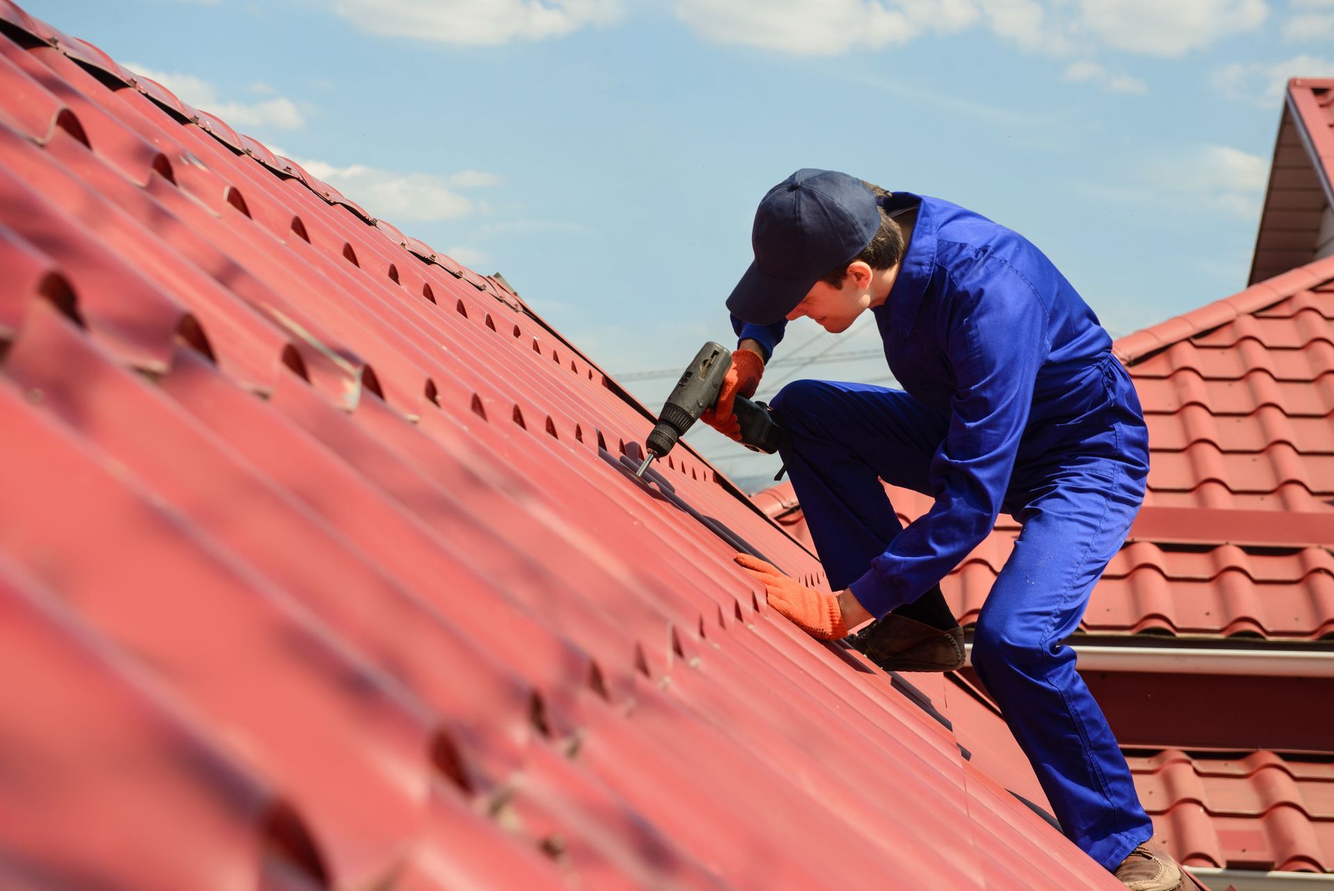 A roofer in a blue uniform is drilling a red metal roof.