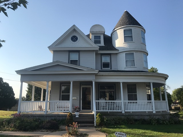 Two-story light-colored house with a wrap-around porch and a turret. Blue sky background.