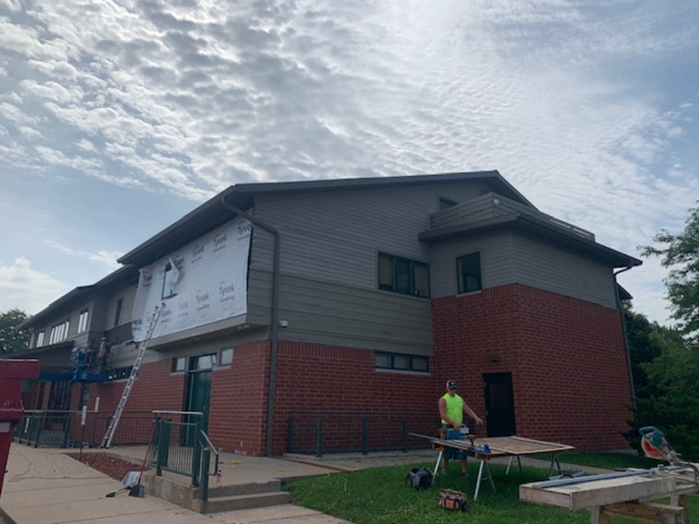 Construction on a two-story building with red brick and gray siding. A worker cuts wood outside on a sunny day.