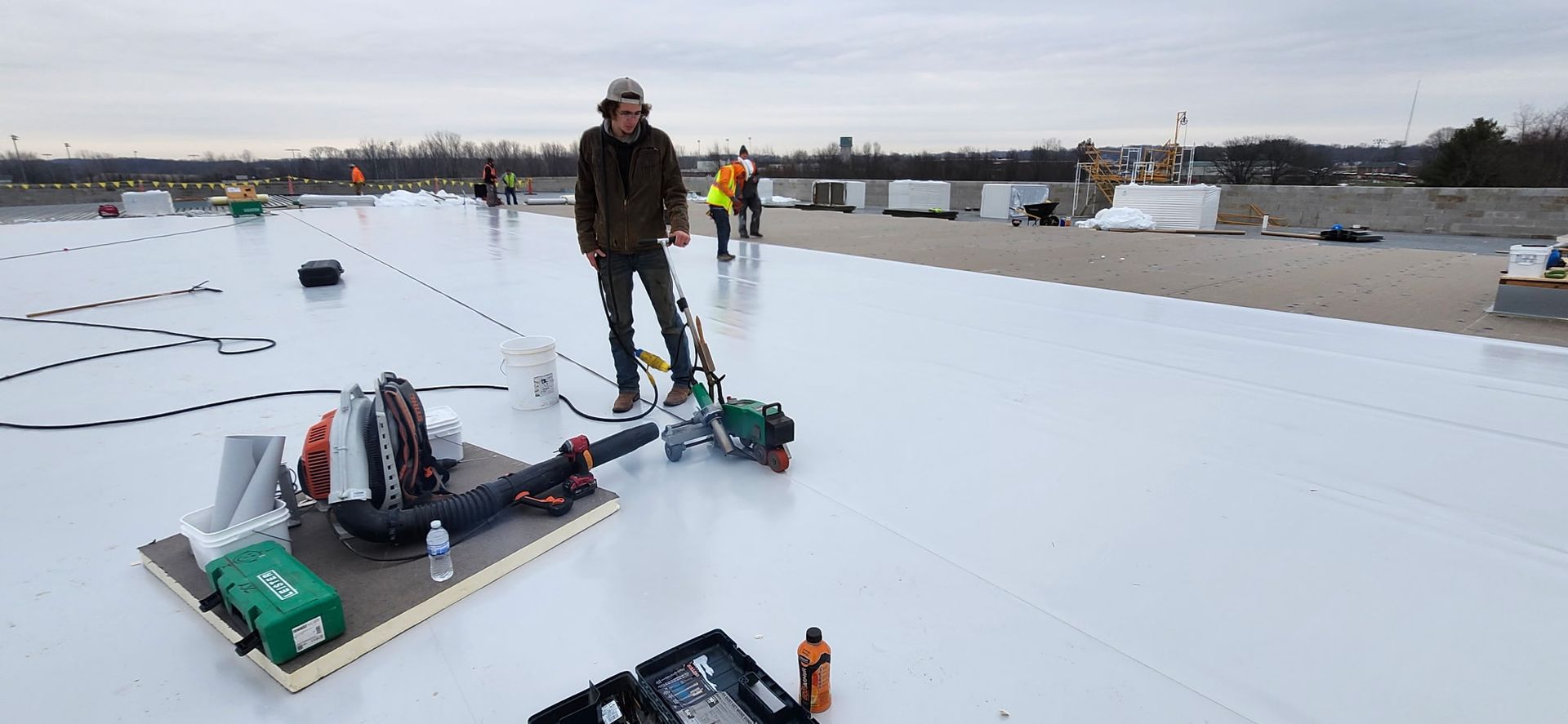 Man using a welding machine on a white rooftop. Construction equipment and another worker in the background. Cloudy sky.
