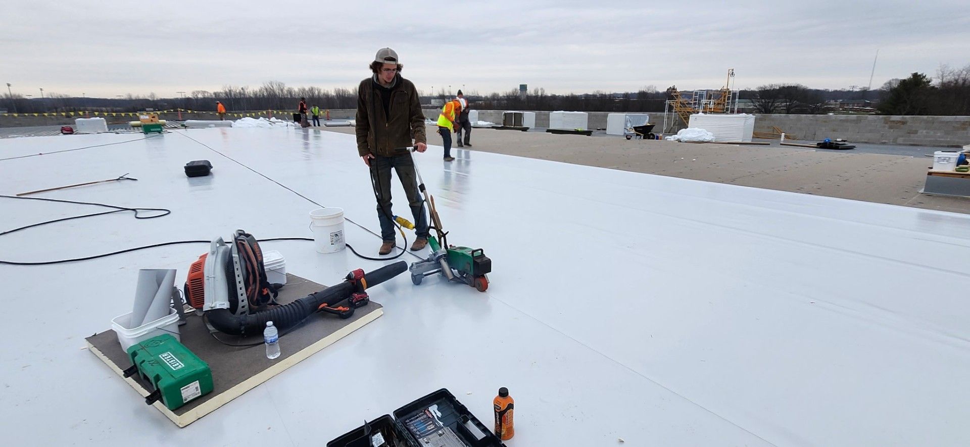 A construction worker welds a white roof membrane on a building rooftop. Other workers are visible in the distance.