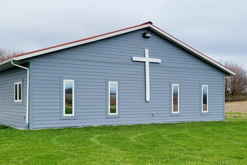 Blue-sided church building with a white cross, rectangular windows, and a red roof on a green lawn.