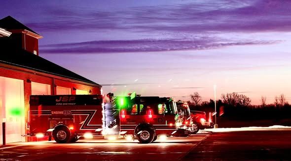 Fire trucks with flashing lights outside a fire station at dusk.