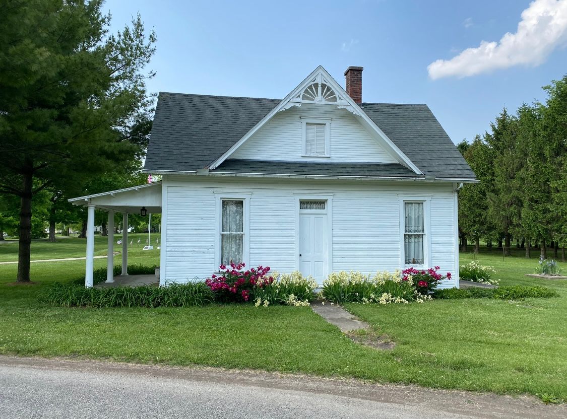 White cottage with porch and ornate gable, surrounded by greenery and flowers.