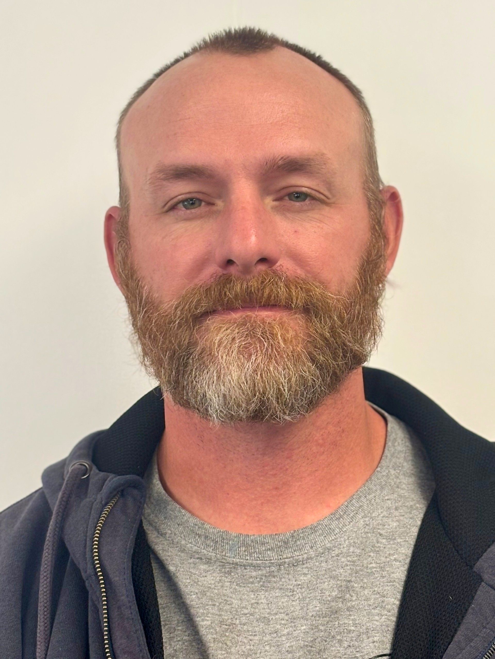 Man with a full beard and short hair, wearing a gray t-shirt and dark jacket, looking directly at the camera.