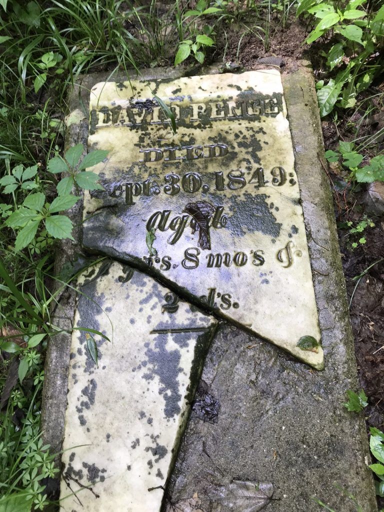 Broken, weathered tombstone in a forest. Partially legible inscription.  Damp, overgrown with foliage.