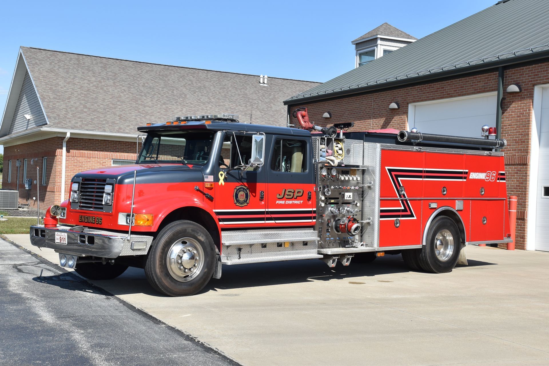 Red and black fire truck parked in front of a brick fire station on a sunny day.