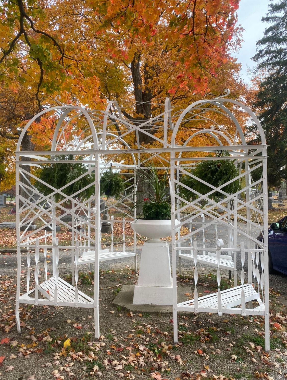 White ornate gazebo with built-in benches, centered on a pedestal, set in a cemetery with autumn foliage.