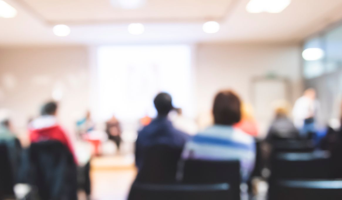 Audience seated in a conference room; blurry view of a presentation in progress on a screen.