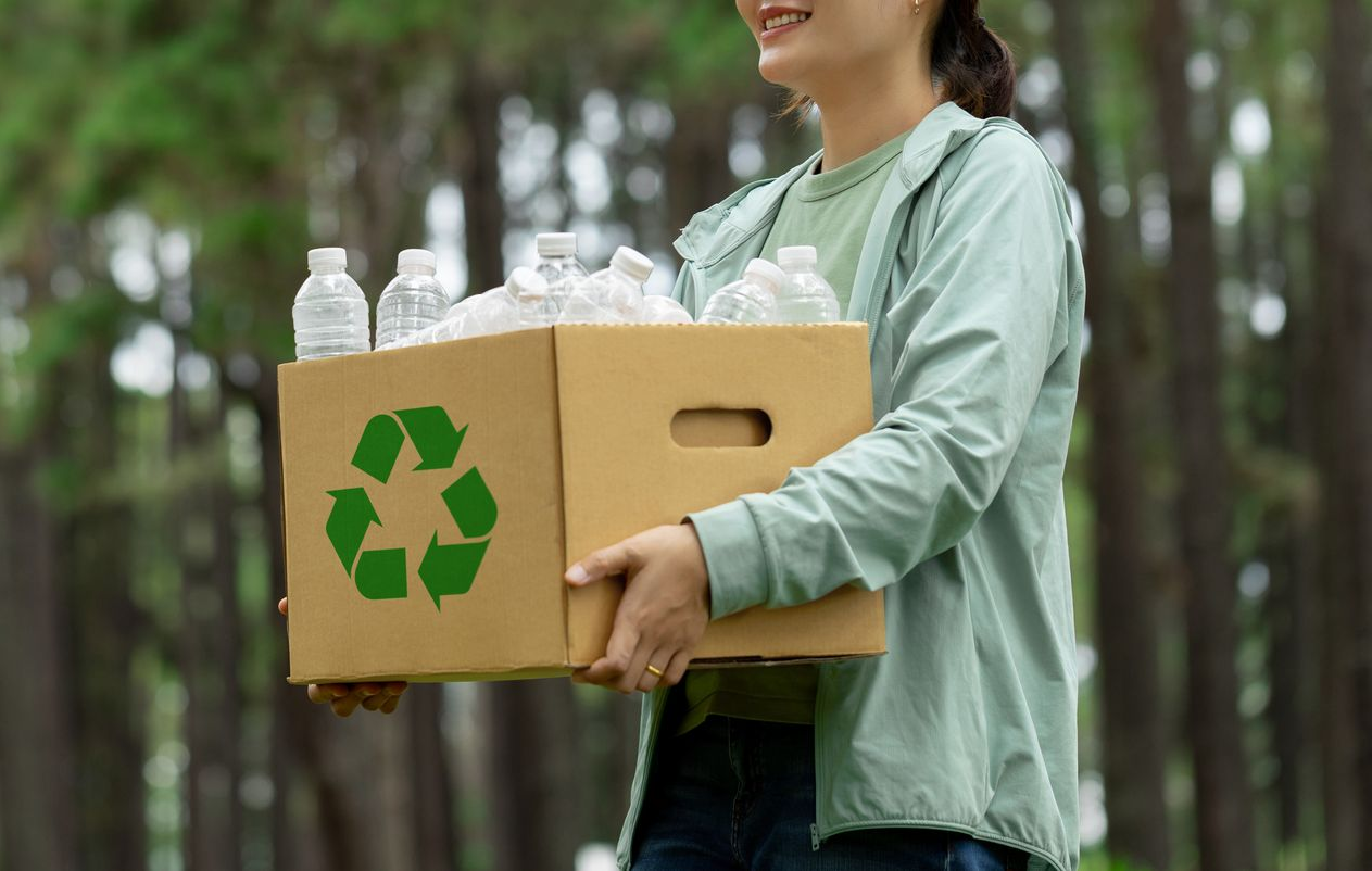Person carrying a cardboard box filled with plastic bottles, recycling symbol on the side, in a forest setting.