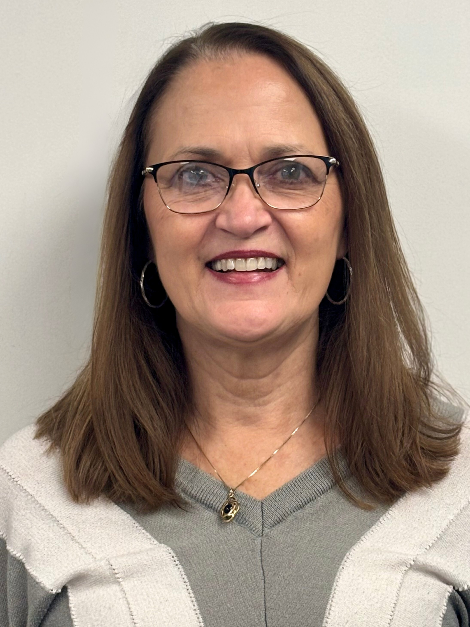 Woman with glasses, smiling, wearing a gray sweater and a necklace, in front of a white wall.