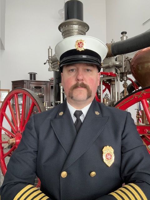 Firefighter in uniform and hat stands in front of antique fire engine.