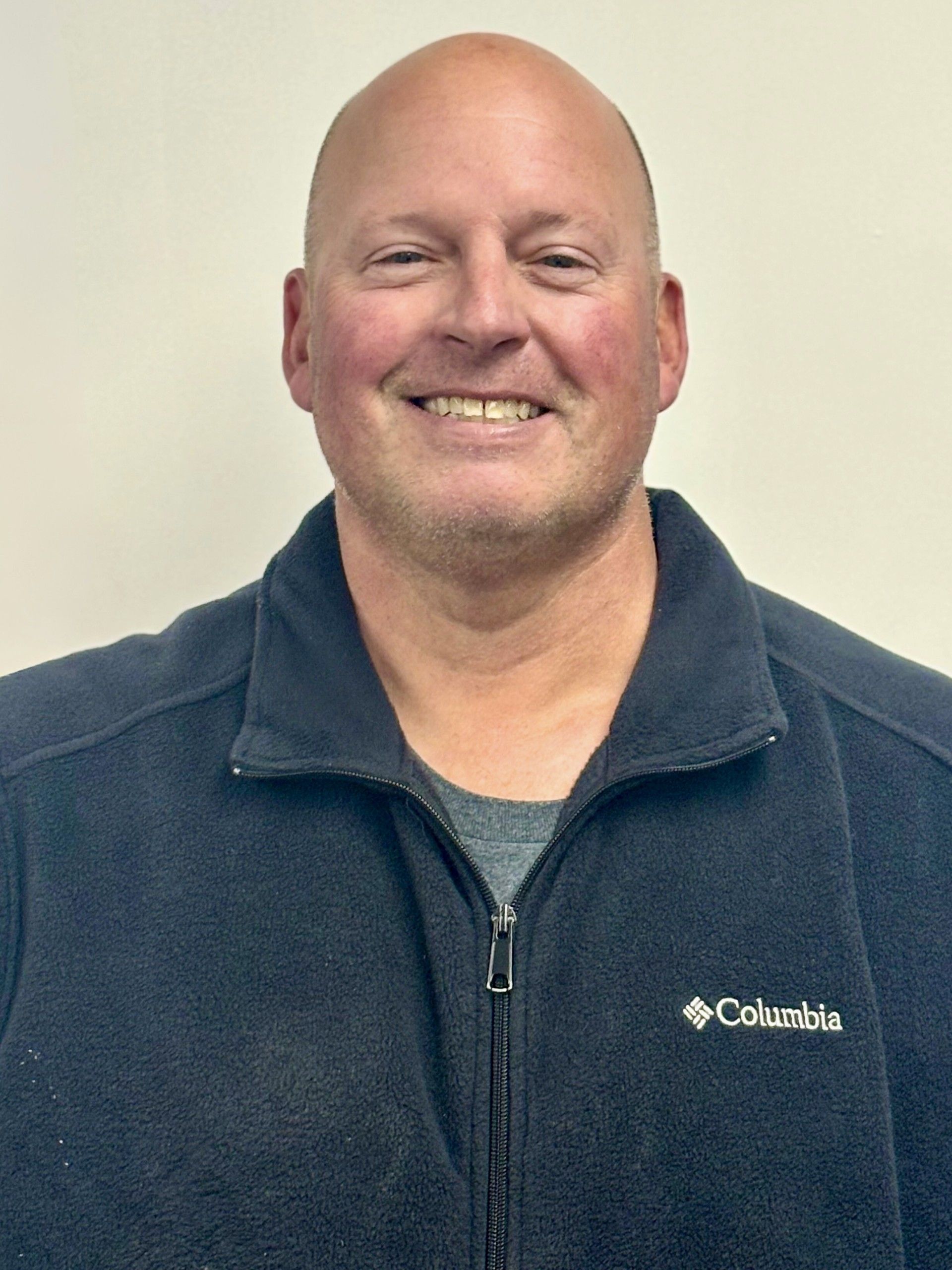 Bald man smiling, wearing a dark blue Columbia fleece jacket in front of a neutral background.