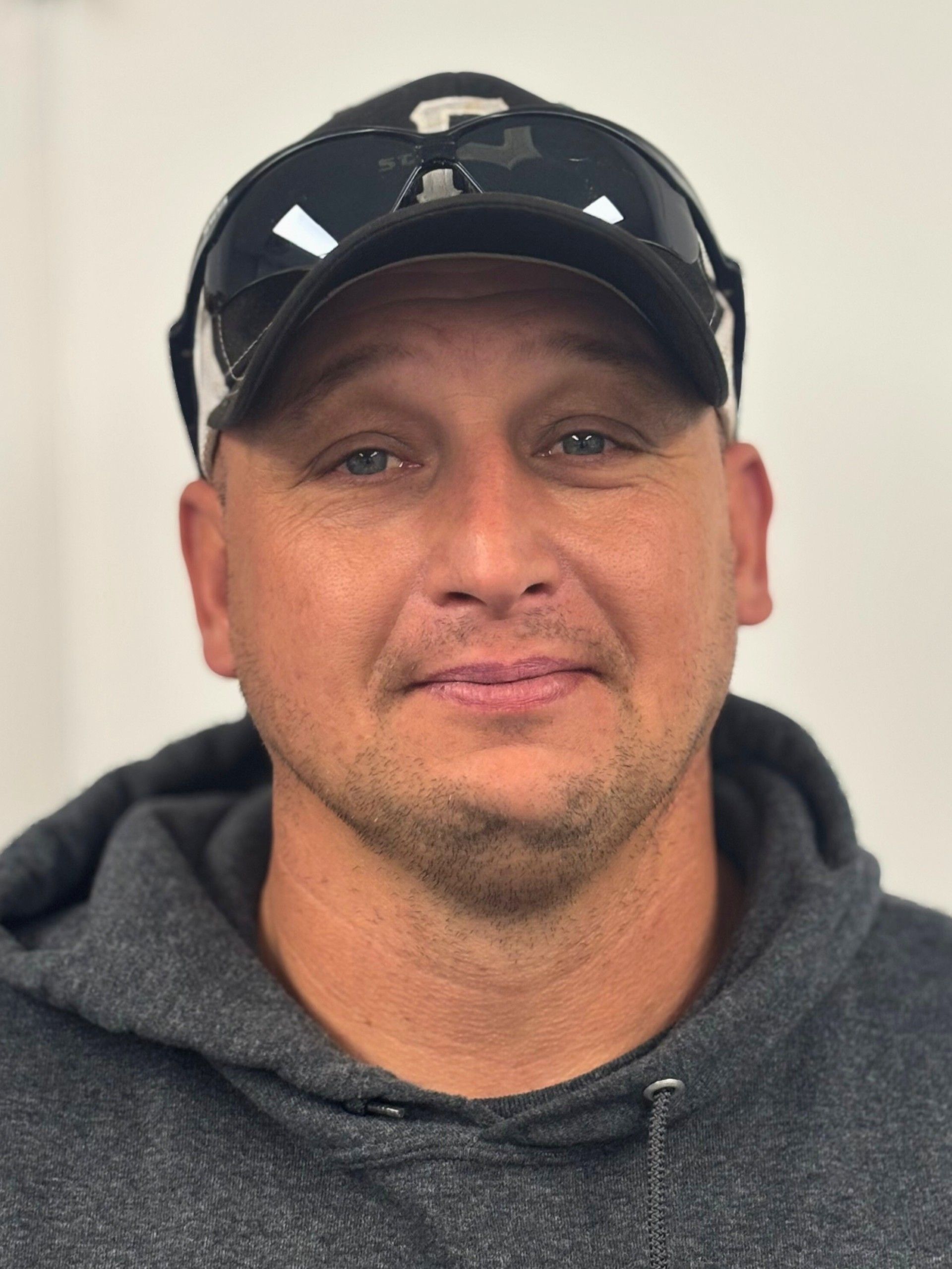 Man wearing a hat and sunglasses, smiling, in a gray hoodie.