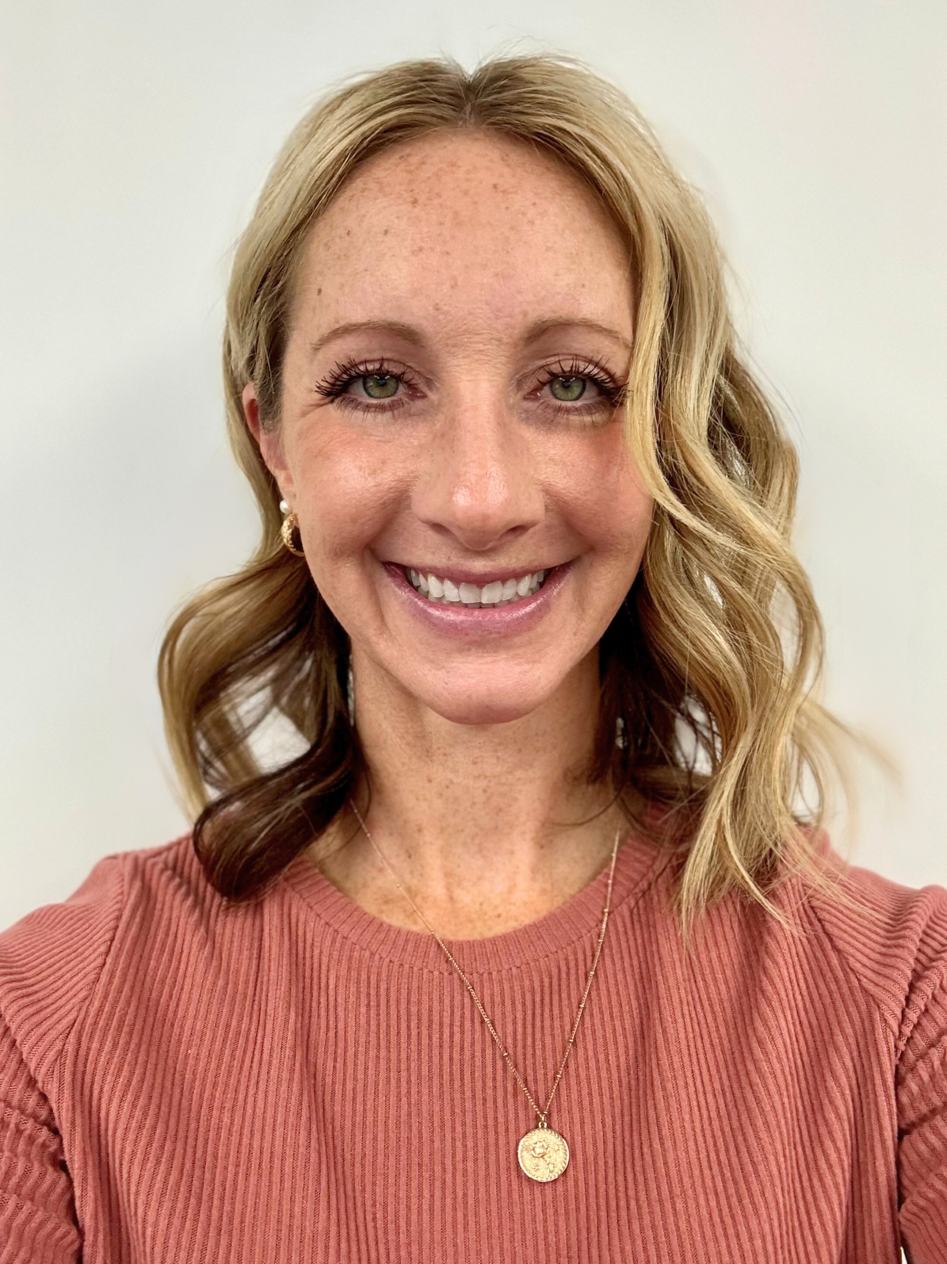 Woman with blonde wavy hair smiles at the camera, wearing a pink sweater and necklace.