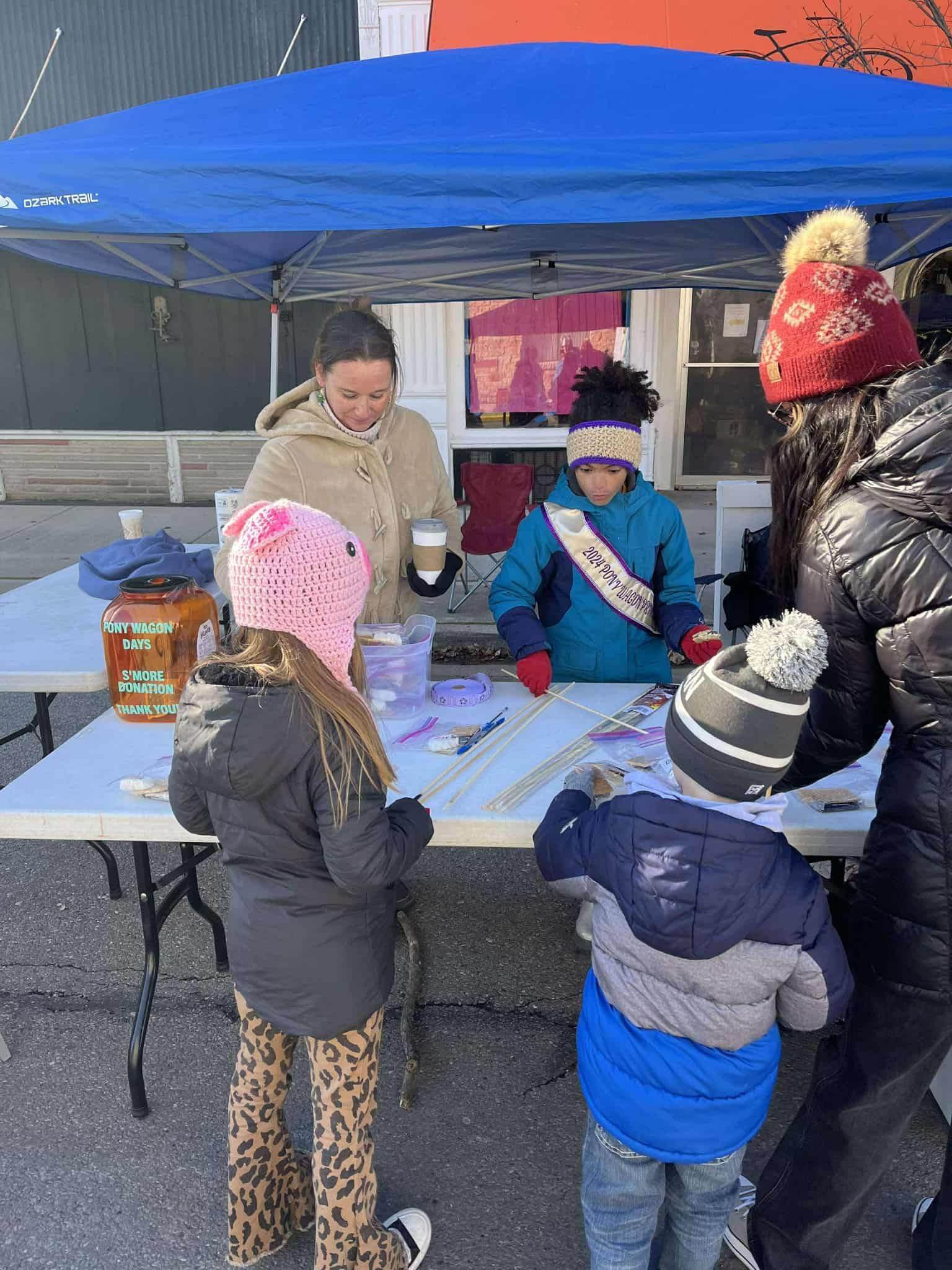 People at a vendor table under a blue tent; items for sale. Children browse jewelry, woman assists, sunny day.