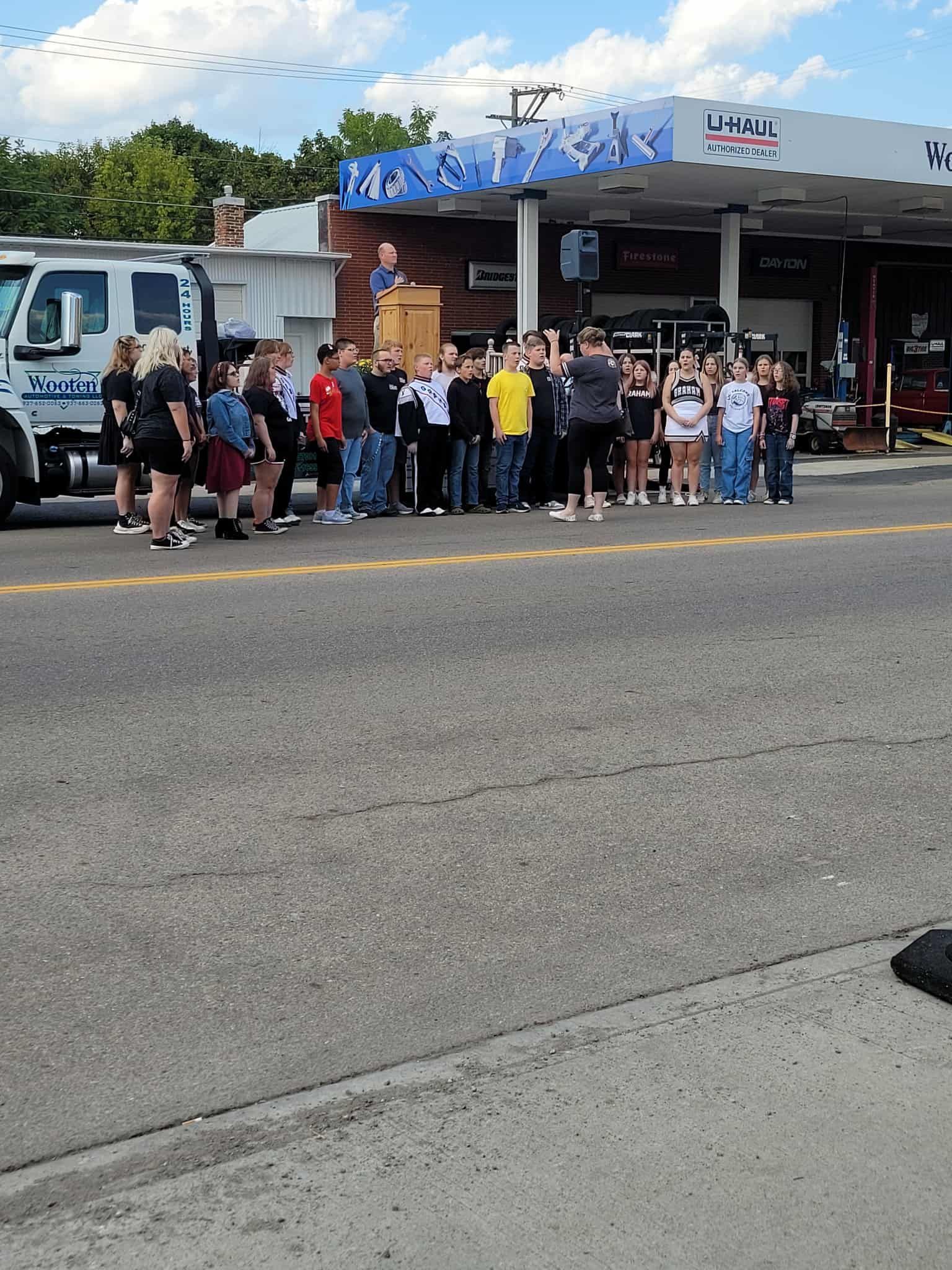Group of people gathered outside a gas station, some looking toward a speaker.