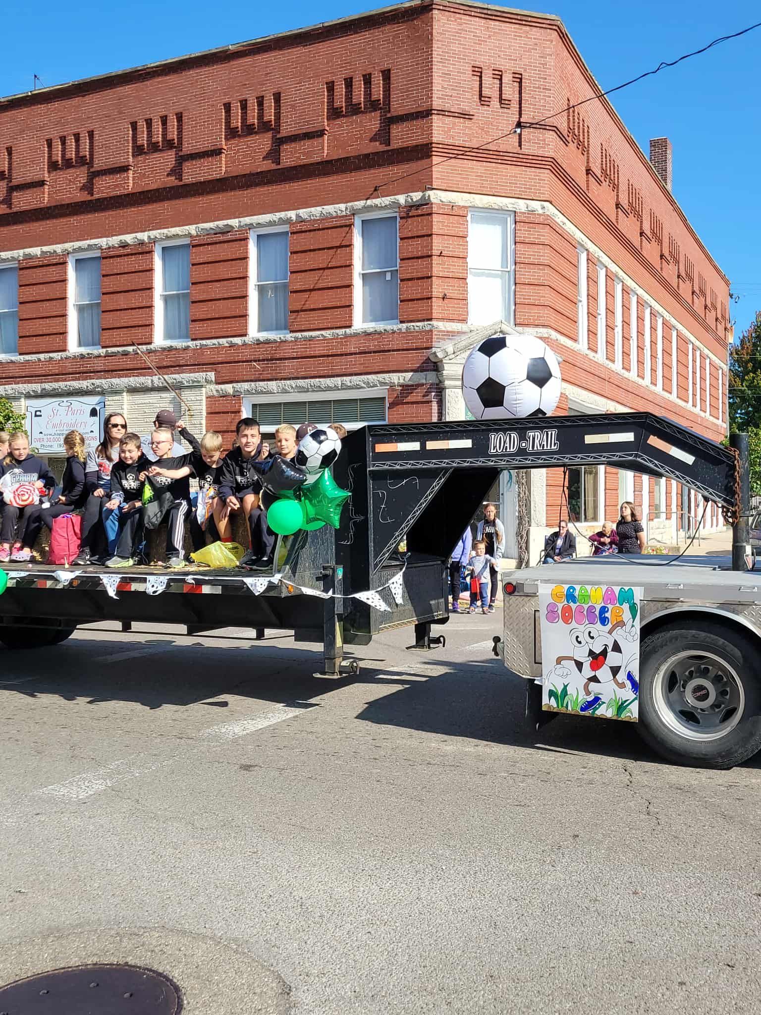 Soccer team on a flatbed trailer in a parade; large soccer ball and building in background.