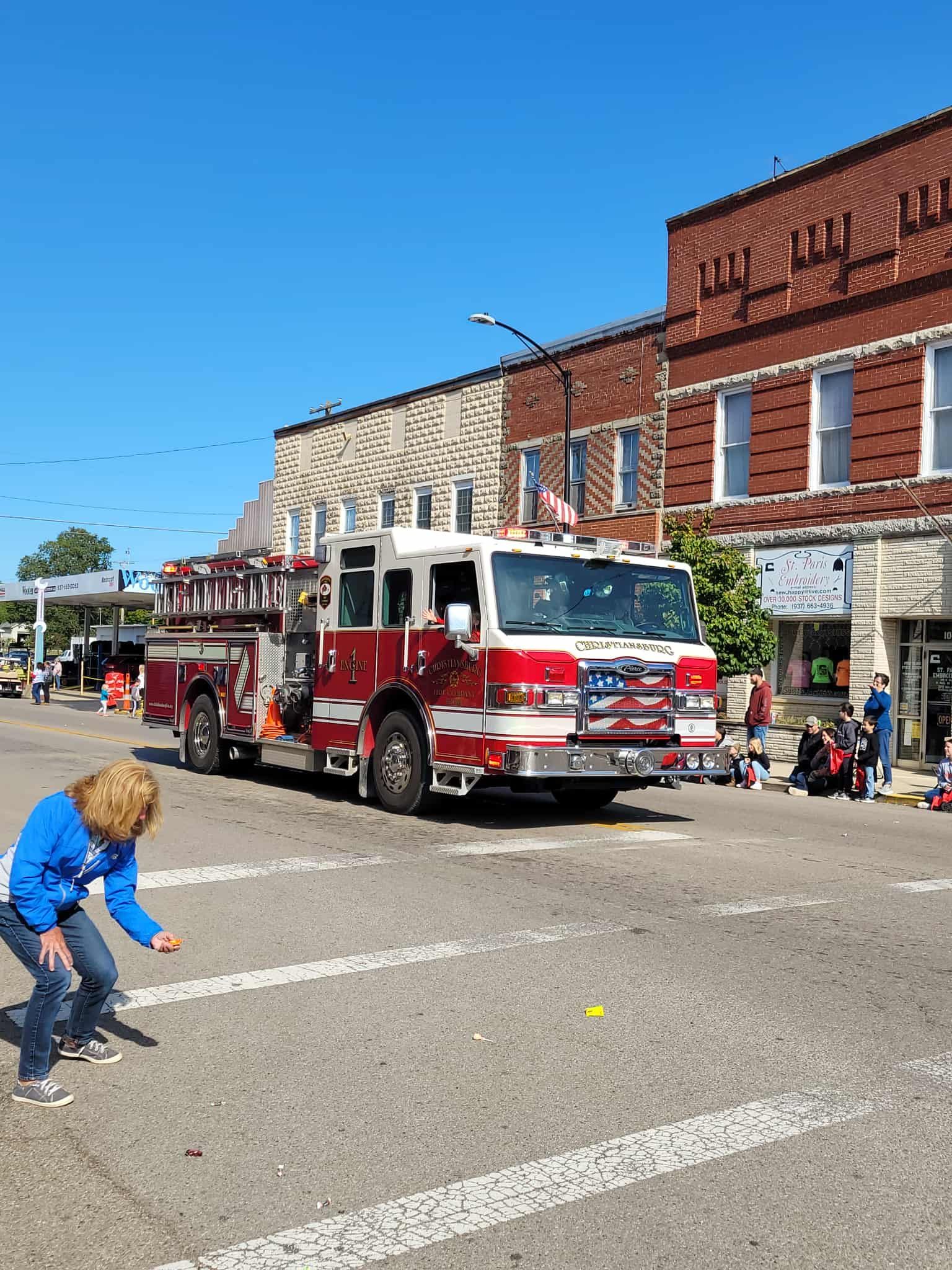 Fire truck in a parade, driving past buildings; a child crouches to pick something up in the street.