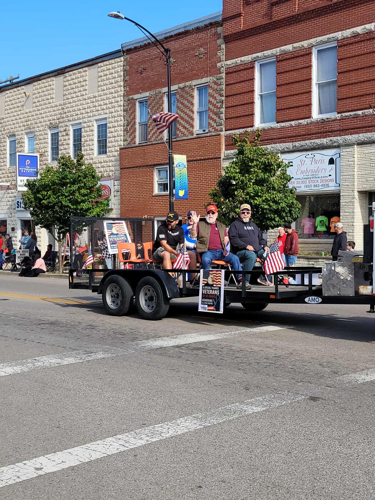 Parade float with people seated, waving flags, in front of downtown buildings.