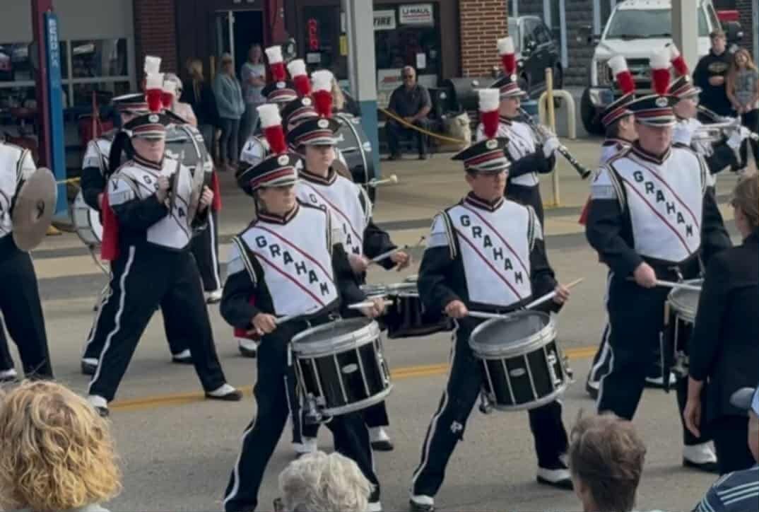 Marching band in black and white uniforms with red accents playing drums in a street parade.