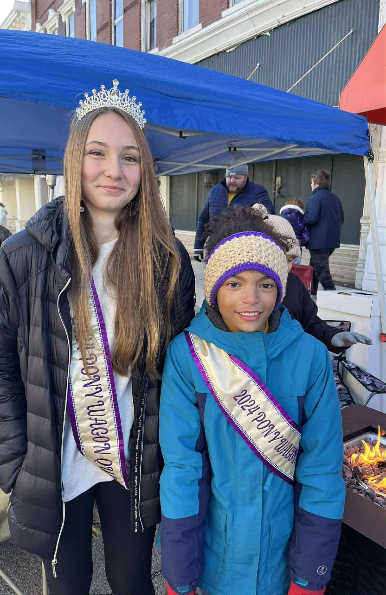 Two girls wearing sashes and one with a tiara pose outdoors. One wears a black jacket, the other a blue coat.