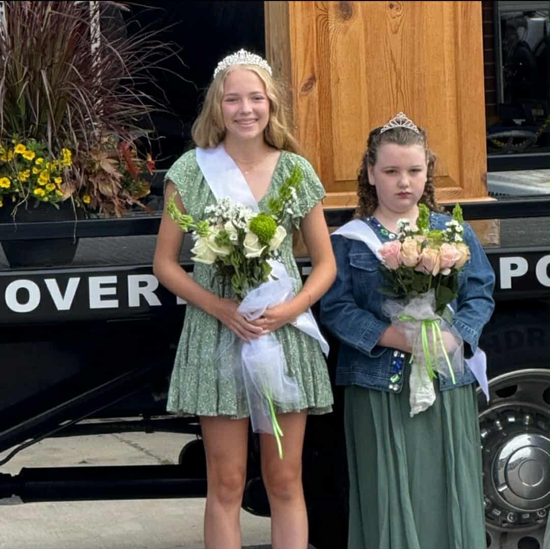 Two girls in crowns and sashes, holding bouquets, stand in front of a vehicle. One in a green dress smiles.