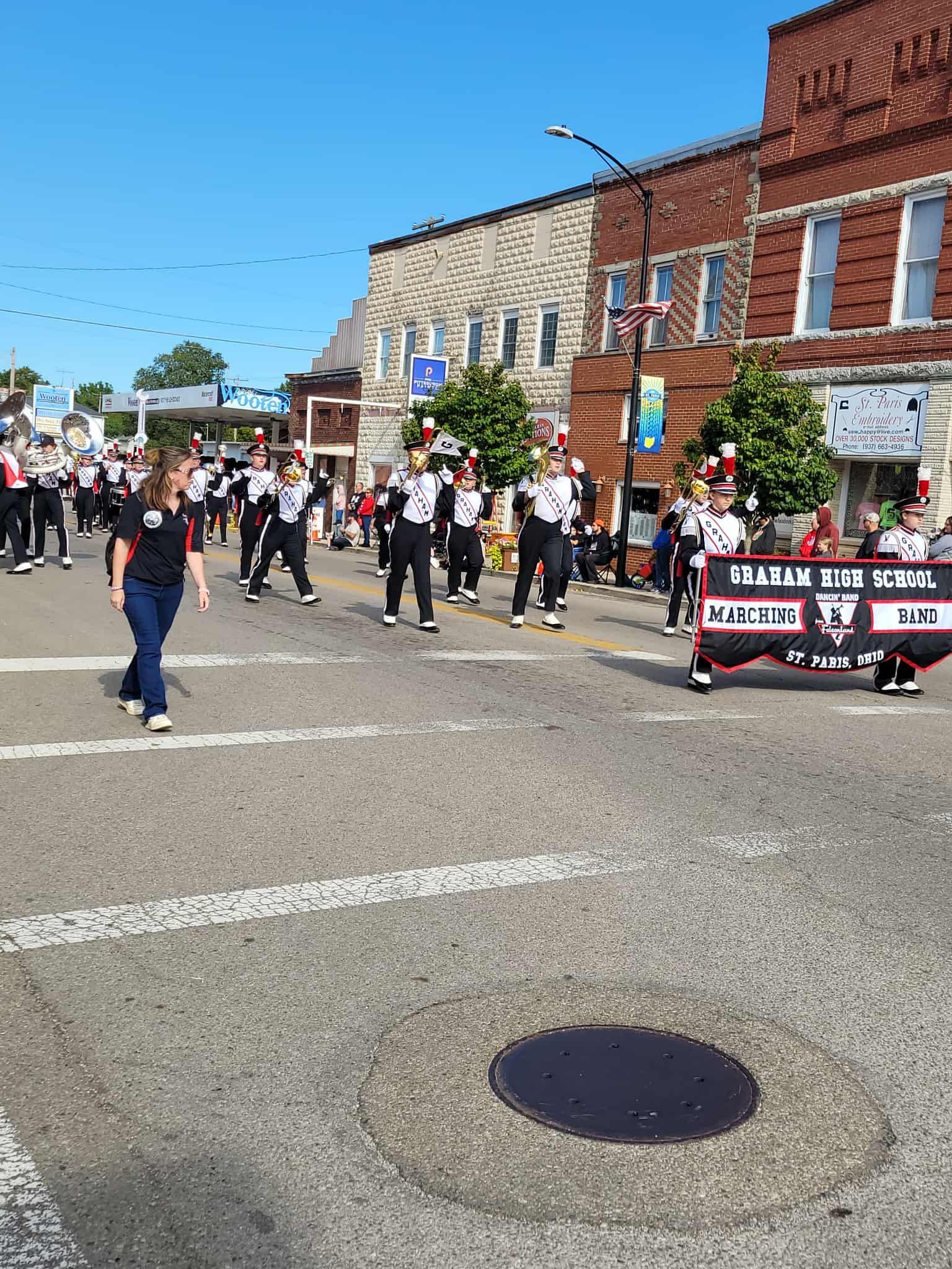 Marching band in parade on street, buildings in background. Members wear black and white uniforms.