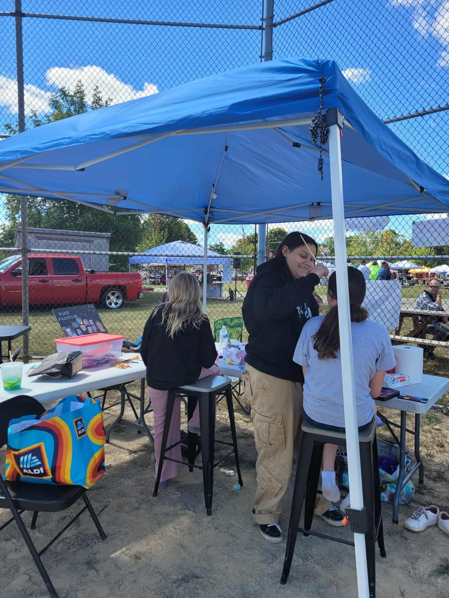 Face painting booth under a blue canopy. A person paints a child's face while another sits at a table.