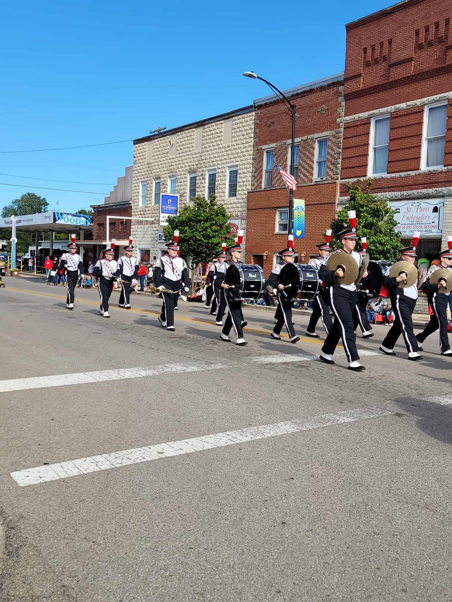 Marching band in black and white uniforms marching down a street in front of brick buildings on a sunny day.
