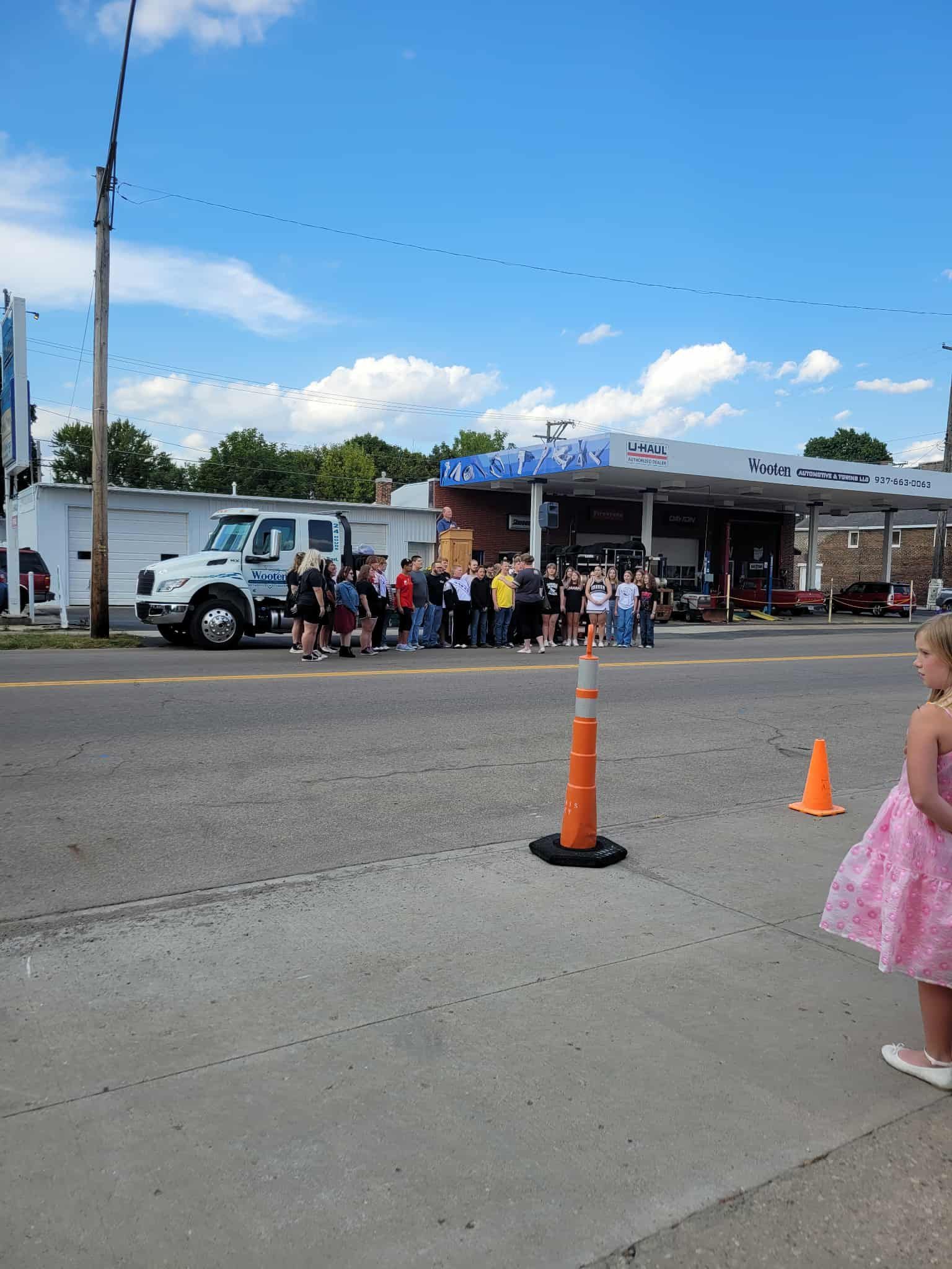 People gathered in front of a gas station under a blue sky, with a white truck and orange traffic cones.