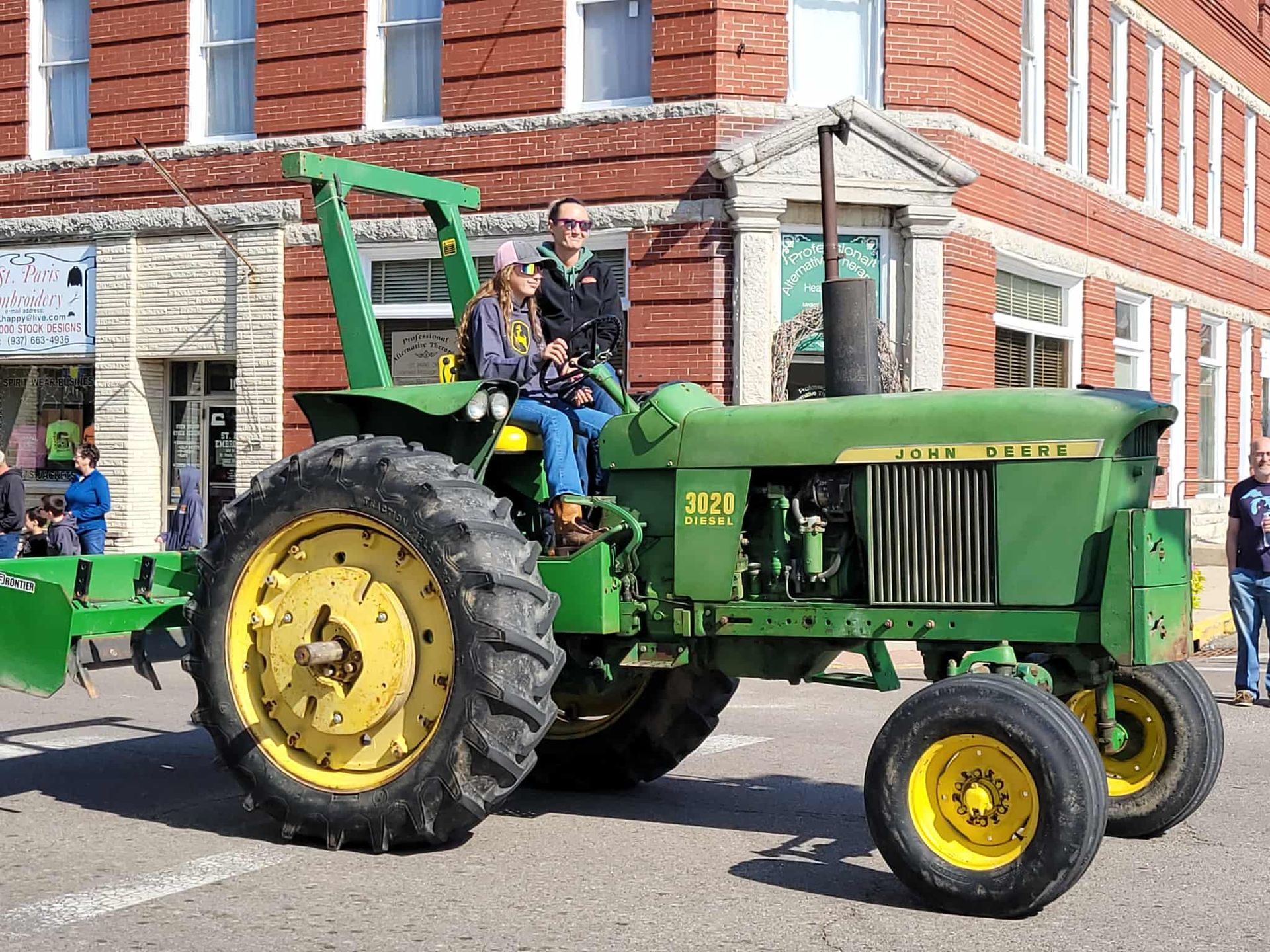Green John Deere tractor with two people driving in a parade, brick buildings in background.