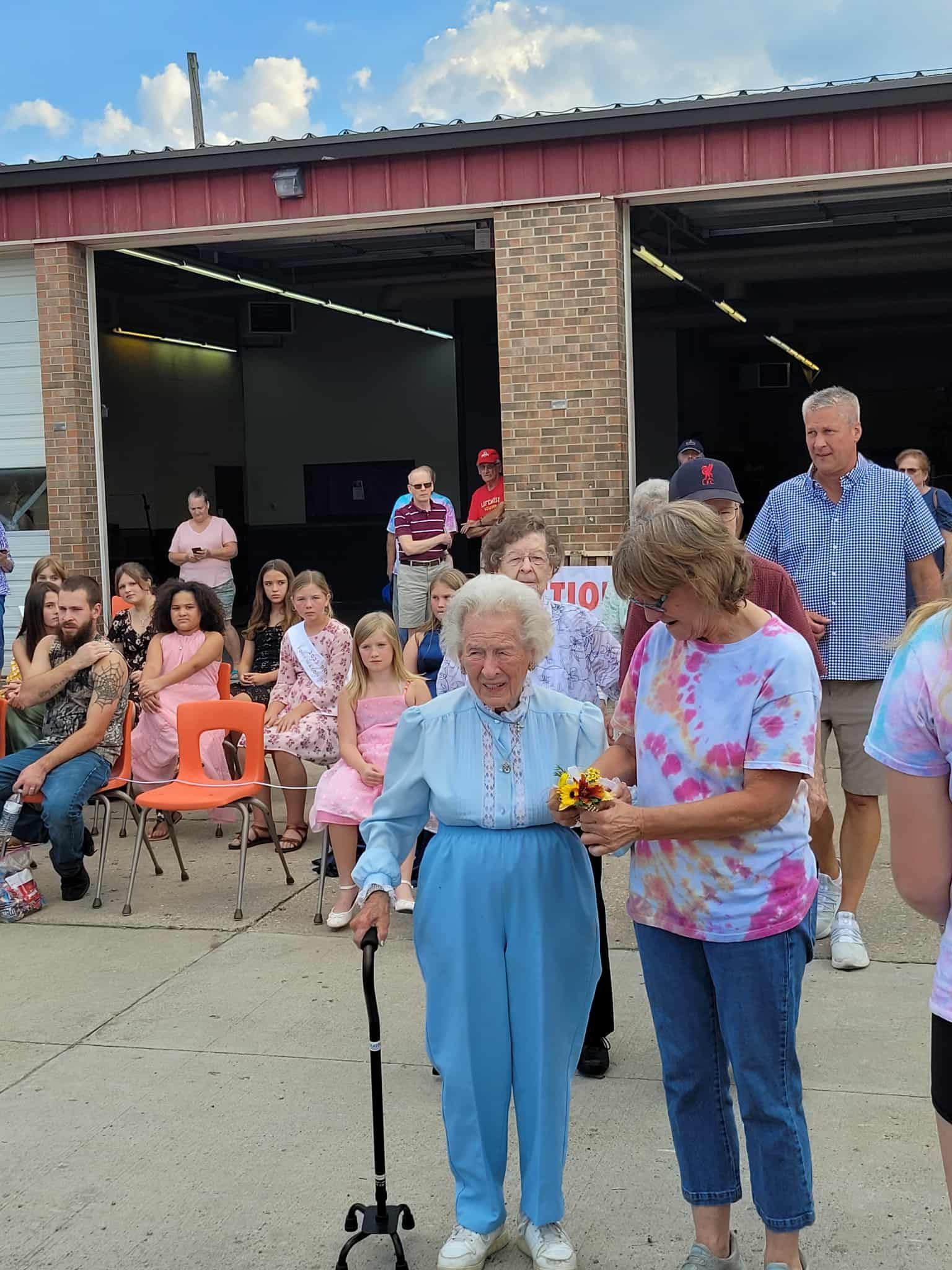 An elderly woman with a cane is presented flowers. A crowd gathers outside a garage with open doors.