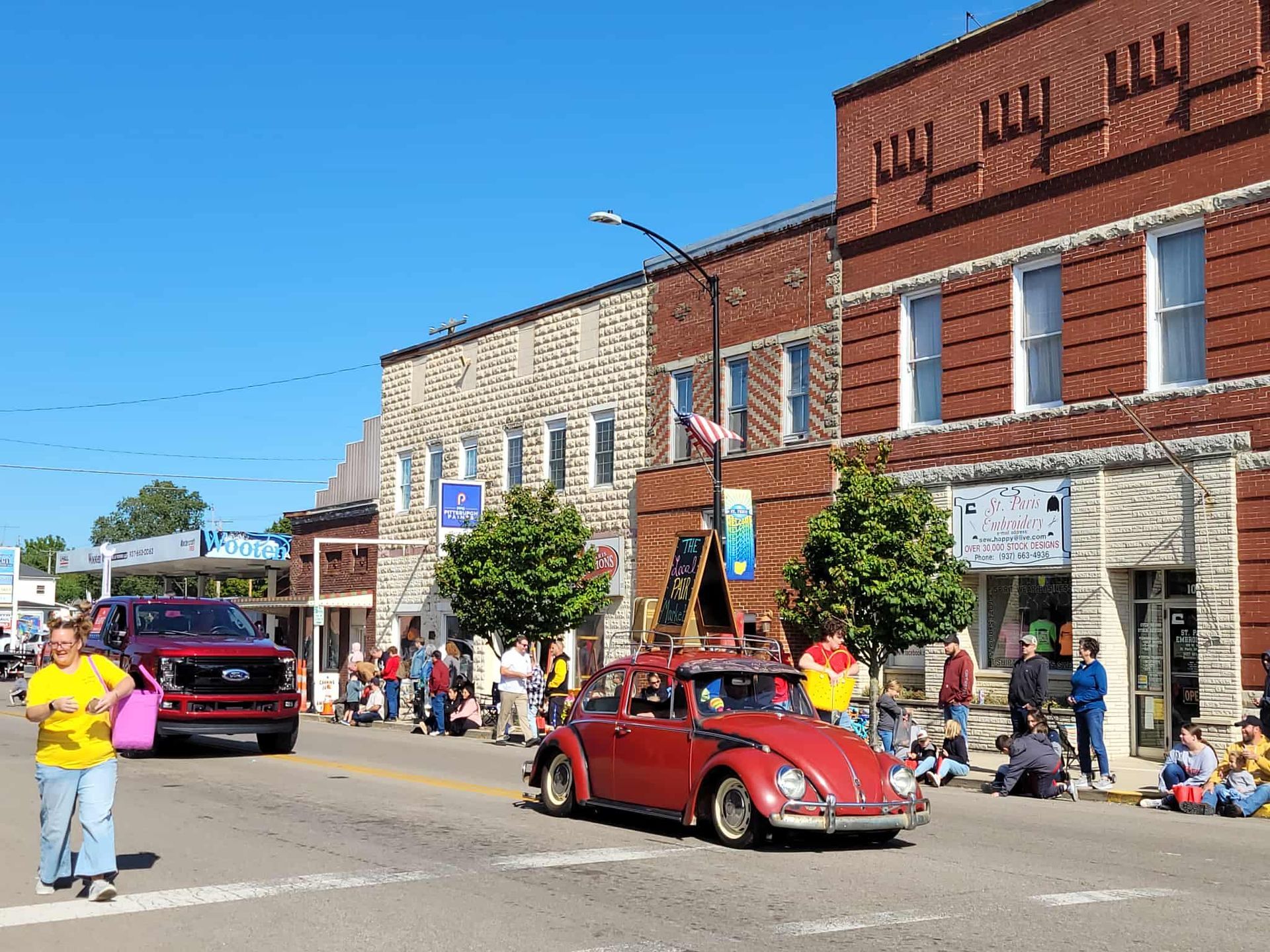 Parade on Main Street with red classic car, people watching, and brick buildings under a blue sky.