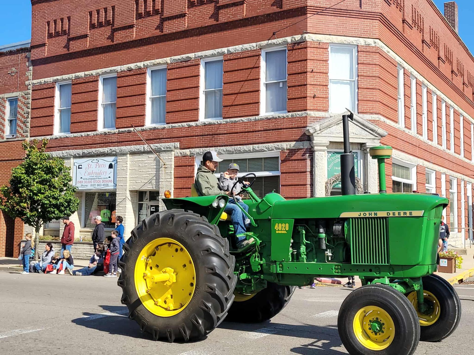 John Deere tractor in a parade, with two people aboard, driving past a brick building.