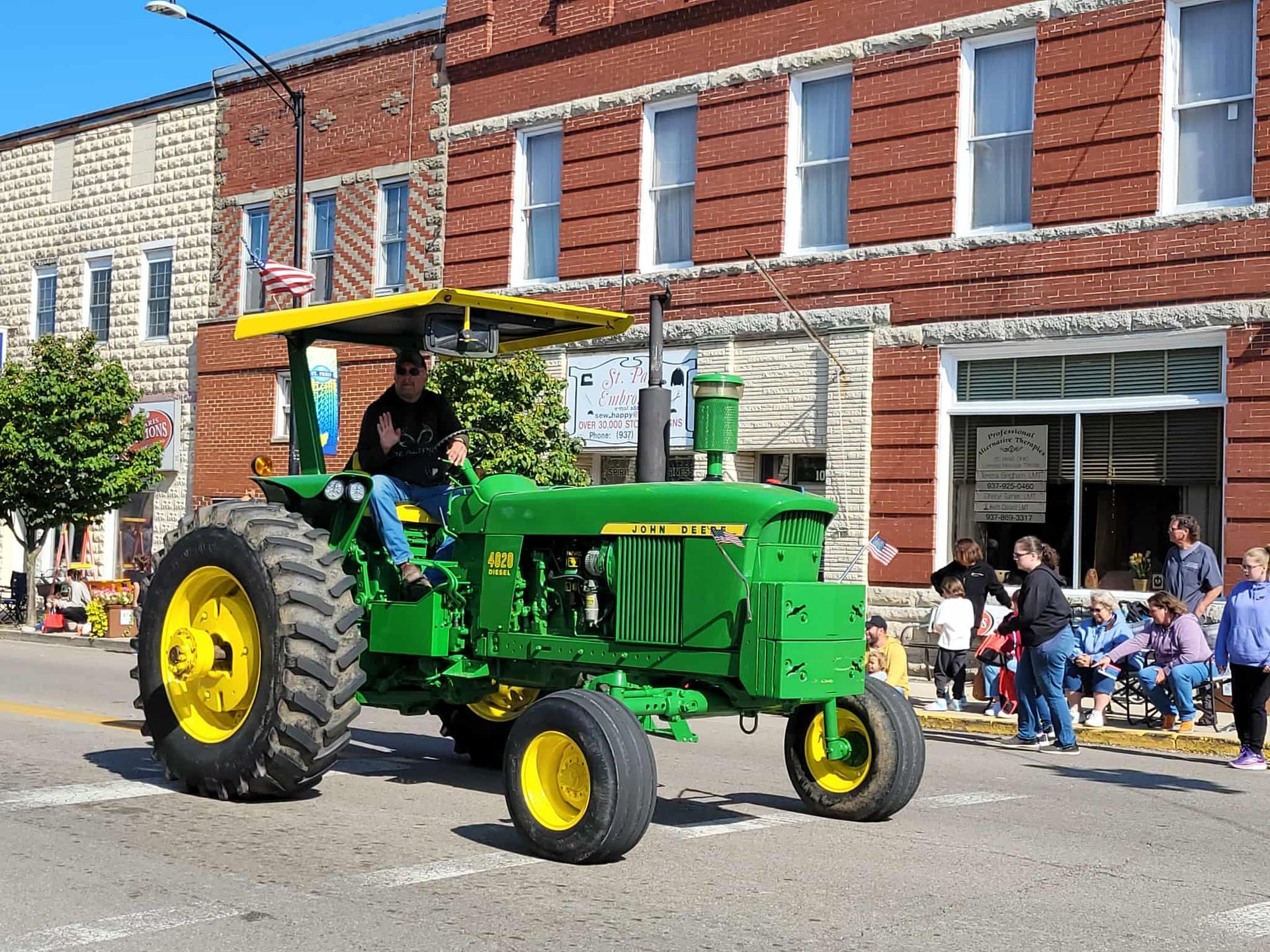 Green John Deere tractor in a parade, driver waving, buildings in background.