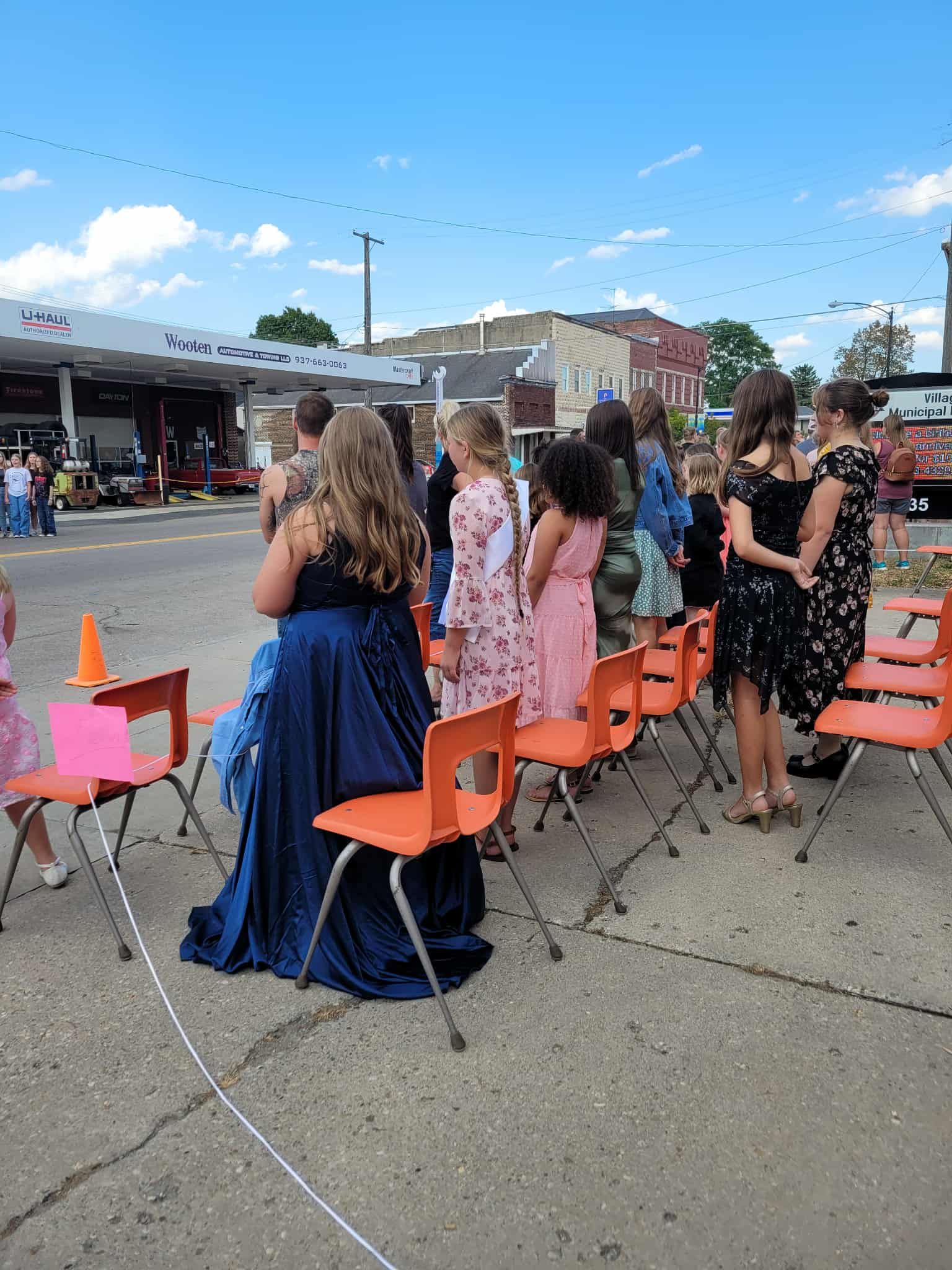 Group of people in formal attire standing in a row of orange chairs outside a gas station on a sunny day.