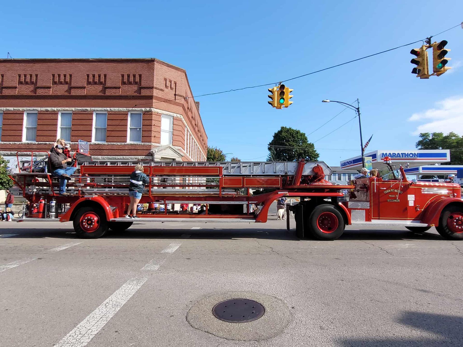 Vintage red fire truck on a street, two people on the ladder, brick building in the background.